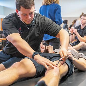 Male student practices listening to blood flow on another student’s leg.